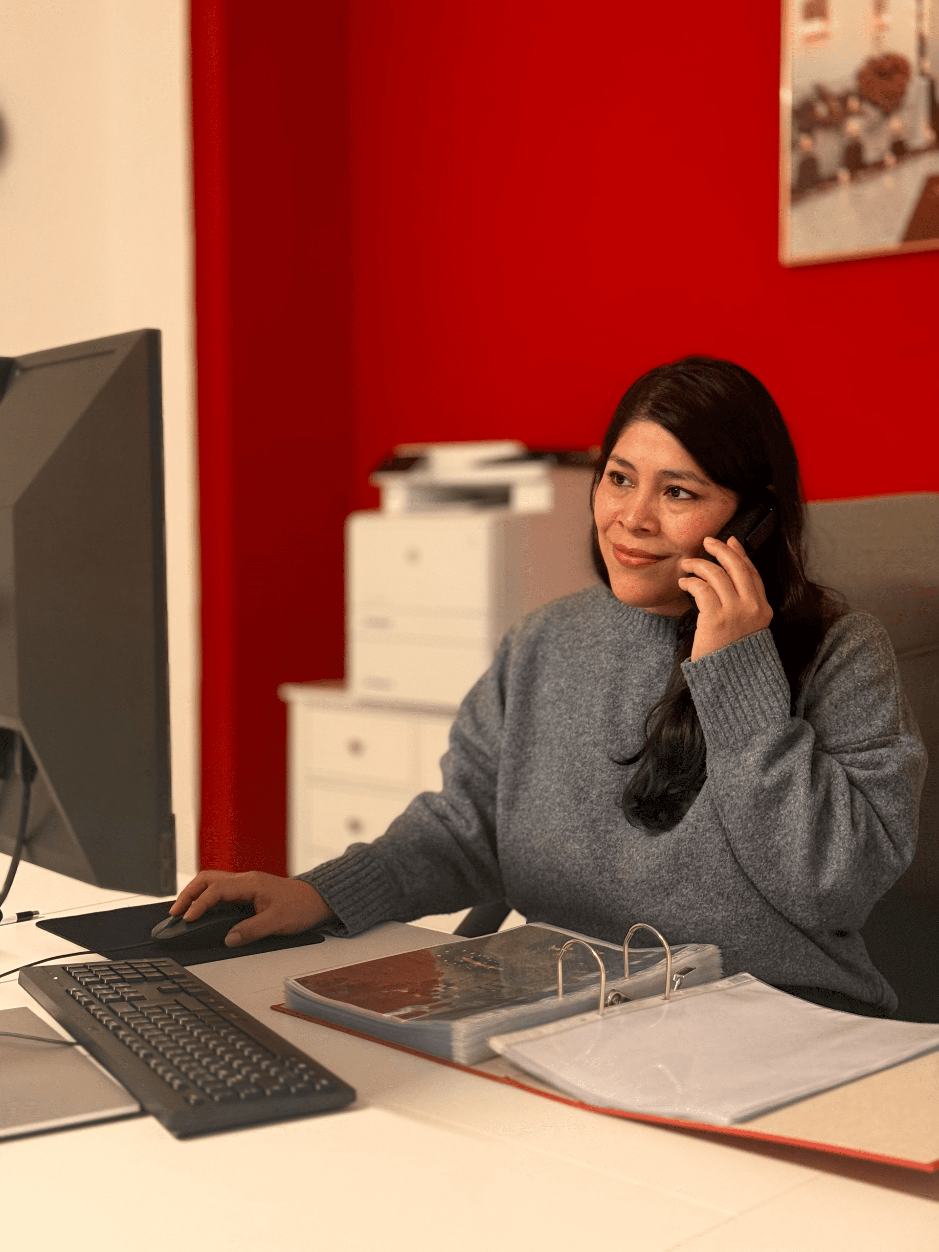 Eine Frau sitzt an einem Schreibtisch, benutzt einen Computer und spricht am Telefon. Sie trägt einen grauen Pullover und hat einen offenen Ordner, eine Tastatur und eine Maus vor sich. Der Hintergrund ist rot und weiß mit sichtbaren Bürogeräten.